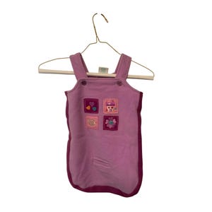 May include: A lavender baby overall with a dark pink trim and four embroidered patches. The patches feature hearts, a house, a flower, and a bear. The overall is hanging on a gold hanger.