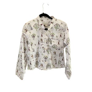 Pode incluir: Uma camisa branca de manga comprida com botões e uma estampa caprichosa de pessoas e flores. A camisa tem uma gola mandarim e um único bolso no peito esquerdo.