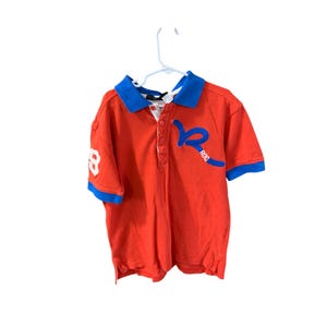 May include: Red short-sleeved polo shirt with a blue collar and trim. The shirt has a white "ROC" logo on the front and a white "8" on the left sleeve.
