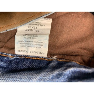 May include: A close-up of a blue denim jean label with white text. The label reads "RN130273 A55394", "MADE IN BANGLADESH", "HECHO EN BANGLADESH", "FABRIQUE AU BANGLADESH", "100% COTTON", "100% ALGODON", "COTON", "33LTDDR", "29X32", "OPD-10H WWMH", and "755107".