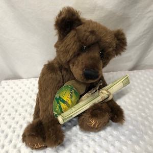 May include: A brown teddy bear holding a rolled-up scroll with a gold ribbon. The bear has a tag that says "Into the Millennium The Bear"