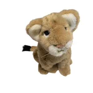 May include: A plush lion cub toy with a light brown body, white-lined ears, and a black-tipped tail. The toy has a soft, textured appearance and is positioned against a white background. The cub's eyes are black, and it has a pink nose.