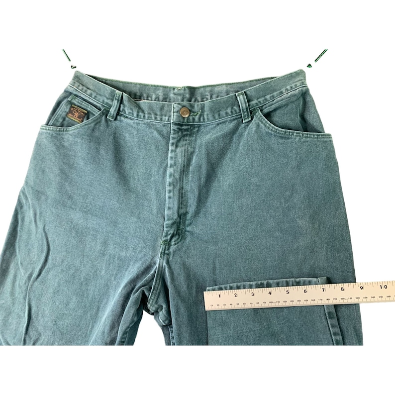 May include: A pair of green denim jeans with a ruler for size reference. The jeans have a straight leg and a classic five-pocket design.