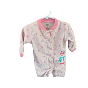 May include: Pink baby onesie with a snowman design and snowflake print. The onesie has a button closure and long sleeves. The text "Carter's" is printed on the onesie.