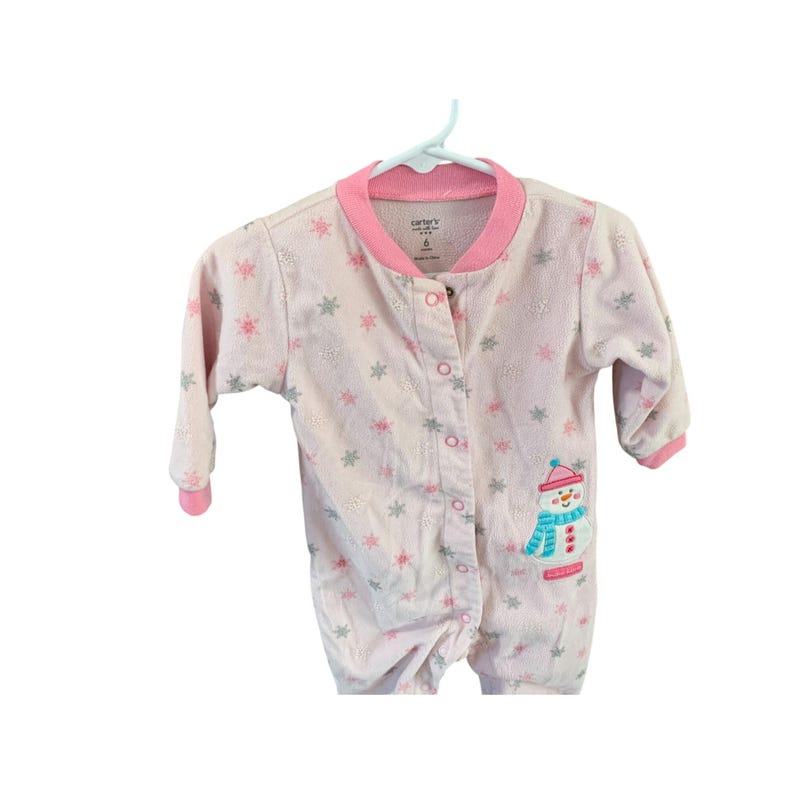 May include: Pink baby onesie with a snowman design and snowflake print. The onesie has a button closure and long sleeves. The text "Carter's" is printed on the onesie.