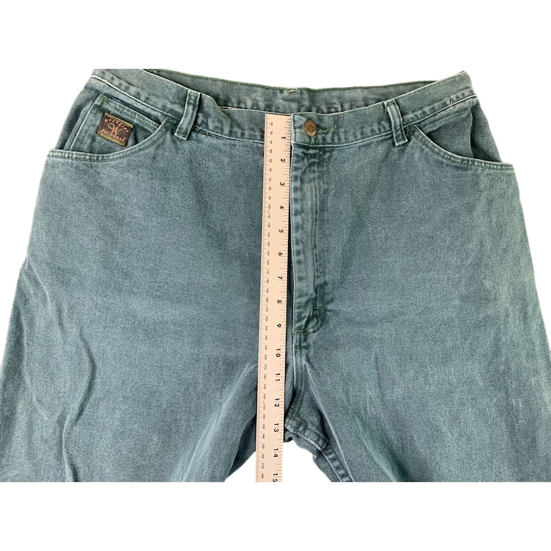 May include: A pair of green denim jeans with a brown leather patch on the back pocket. The jeans are laid flat and a ruler is placed along the side seam to show the length.