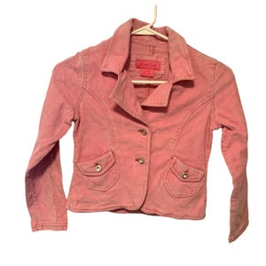 May include: A light pink corduroy blazer with a classic collar, two front pockets, and three buttons. The jacket is hanging on a white hanger. The label reads "Angelina".