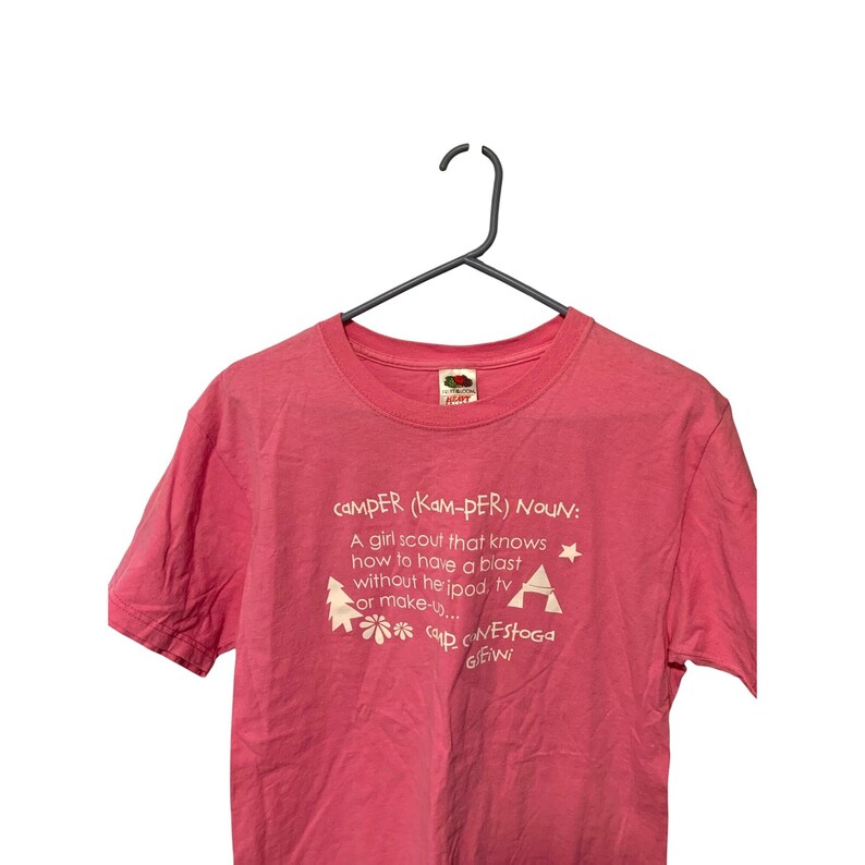K&ouml;nnte beinhalten: Rosafarbenes T-Shirt mit wei&szlig;em Text und Grafiken. Der Text enth&auml;lt das Wort &bdquo;CAMPER&ldquo; und eine Definition. Grafiken: Baum, Stern und Blume. Das T-Shirt h&auml;ngt an einem schwarzen Kleiderb&uuml;gel.