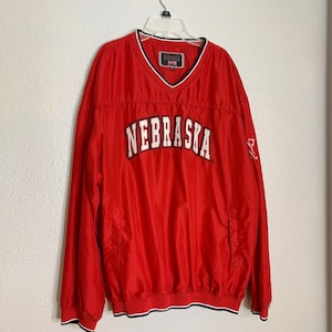 以下が含まれることがあります： 赤いウィンドブレーカーのジャケットで、前面に白い文字で「NEBRASKA」と書かれています。ジャケットは首と裾に黒と白のトリムが付いています。