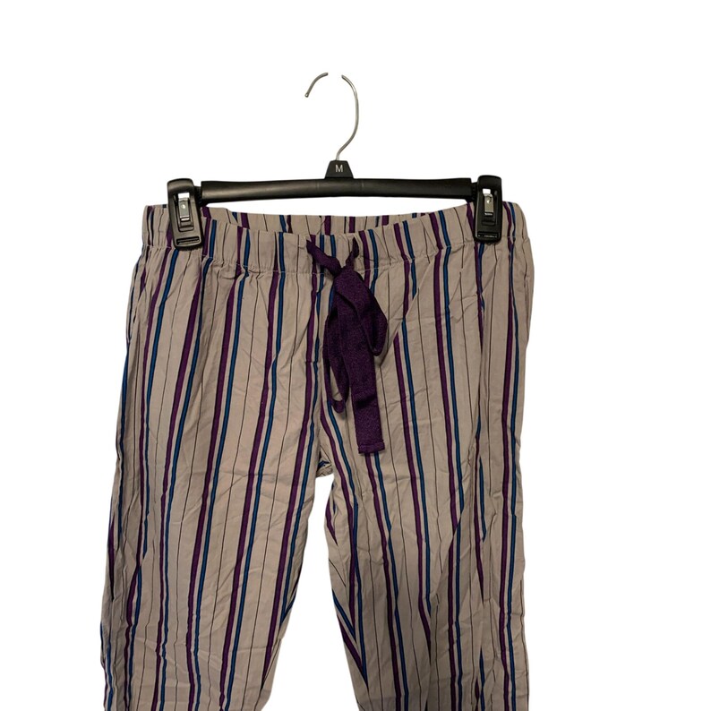 May include: Striped pajama pants in a neutral color with vertical stripes in purple and teal. The pants have a drawstring waist with a purple tie. The pants are hanging on a black hanger against a white background.