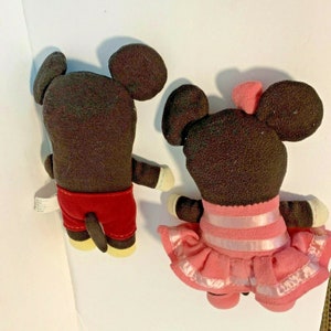 Pu&ograve; includere: Un Mickey Mouse di peluche marrone e una Minnie Mouse di peluche marrone. Mickey indossa pantaloncini rossi e Minnie indossa un vestito rosa con un nastro bianco.