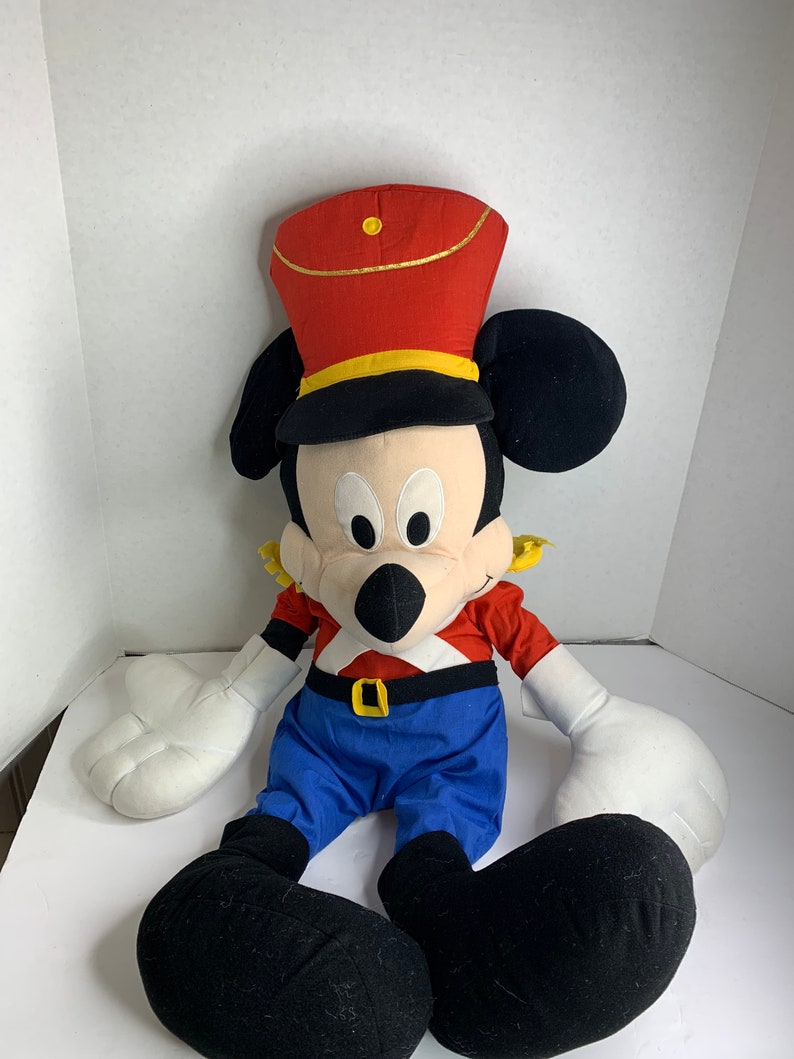 K&ouml;nnte beinhalten: Ein gro&szlig;es Mickey Mouse-Pl&uuml;schtier in einer rot-blauen Marschkapellenuniform. Das Spielzeug hat einen roten Hut mit goldenem Besatz, ein rotes Hemd, blaue Hosen und schwarze Schuhe. Die wei&szlig;en Handschuhe und die gro&szlig;en schwarzen Ohren des Spielzeugs sind ebenfalls sichtbar.