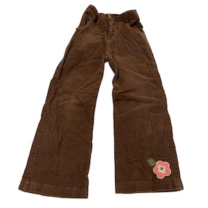 Puede incluir: Pantalón de pana marrón con una flor rosa bordada en la pierna derecha.