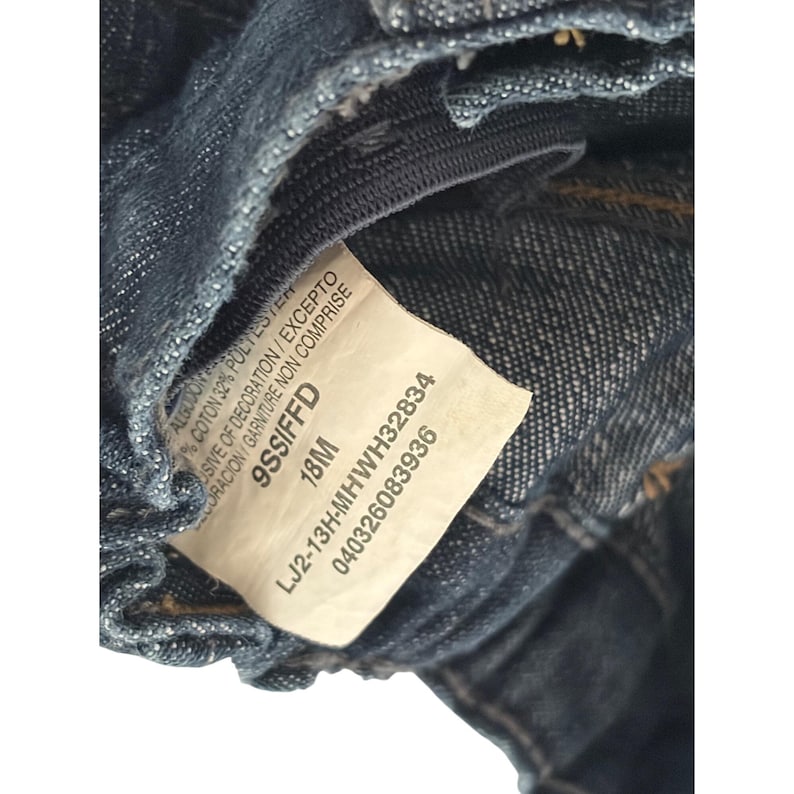 May include: Close-up of dark blue denim fabric with a white tag. The tag has text including "9SSIFFD", "18M", and "LJ2-13H-MHWH32834". The fabric is a blend of cotton and polyester.