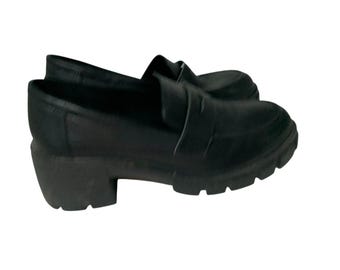 May include: Black leather loafers with a chunky heel. The shoes have a slip-on design with a strap across the top. The soles are thick and have a treaded pattern. These shoes are a stylish and comfortable footwear option.