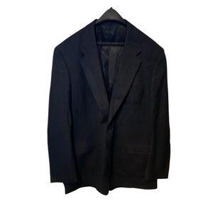 Könnte beinhalten: Ein schwarzer Blazer hängt an einem schwarzen Kleiderbügel. Der Blazer hat ein fallendes Revers, zwei Vordertaschen und einen Ein-Knopf-Verschluss. Der Stoff scheint eine subtile Textur zu haben.