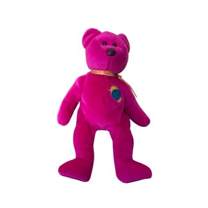 Puede incluir: Un oso de peluche magenta con una cinta dorada alrededor del cuello. El oso tiene un pequeño parche bordado de un globo terráqueo con un sol y el año 1999. El oso tiene una nariz negra y orejas pequeñas y redondas.