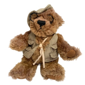 May include: A brown teddy bear wearing a khaki vest and a khaki hat with a pair of round black glasses.