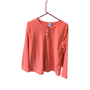 Puede incluir: Una camisa de manga larga de color coral con cuello redondo y tres botones dorados. La camisa está colgada en una percha roja.