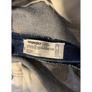 May include: A blue denim Wrangler jeans tag with white text. The tag reads "Wrangler 938DEN 093ADEN 34 X 30" and includes care instructions. The tag also includes the following information: "RN 150273 2000001845 LAR-R434-027 051071500906 73054 MADE IN MEXICO 100% COTTON EXCLUSIVE OF DECORATION"