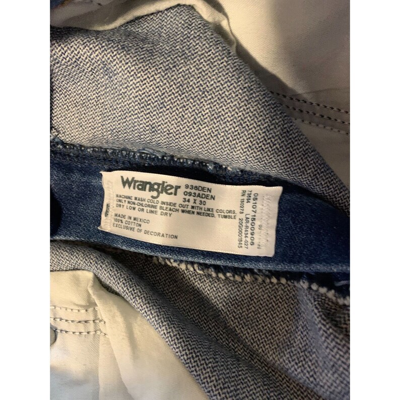 May include: A blue denim Wrangler jeans tag with white text. The tag reads "Wrangler 938DEN 093ADEN 34 X 30" and includes care instructions. The tag also includes the following information: "RN 150273 2000001845 LAR-R434-027 051071500906 73054 MADE IN MEXICO 100% COTTON EXCLUSIVE OF DECORATION"