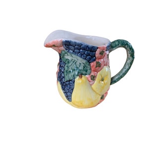 May include: A small ceramic creamer pitcher with a handle, decorated with a colorful fruit design. The design features pears, grapes, and leaves in shades of green, blue, yellow, pink, and red.