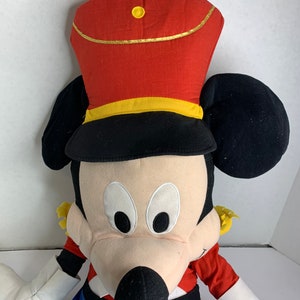 K&ouml;nnte beinhalten: Ein gro&szlig;es Pl&uuml;schtier einer Zeichentrickmausfigur mit roter und schwarzer Uniformm&uuml;tze mit goldenen Verzierungen. Die Maus hat schwarze Ohren, ein cremefarbenes Gesicht und blaue Hosen. Die Arme des Spielzeugs sind wei&szlig;, die Uniform hat rote und gelbe Akzente.