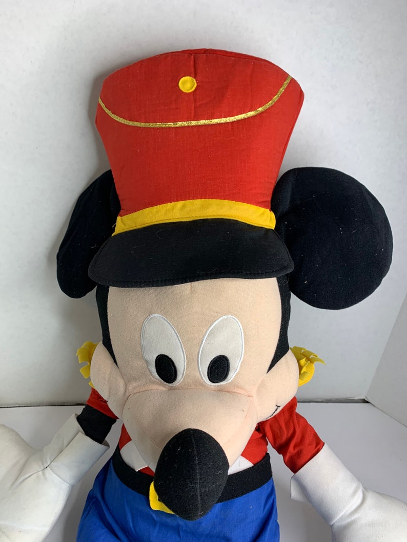 K&ouml;nnte beinhalten: Ein gro&szlig;es Pl&uuml;schtier einer Zeichentrickmausfigur mit roter und schwarzer Uniformm&uuml;tze mit goldenen Verzierungen. Die Maus hat schwarze Ohren, ein cremefarbenes Gesicht und blaue Hosen. Die Arme des Spielzeugs sind wei&szlig;, die Uniform hat rote und gelbe Akzente.