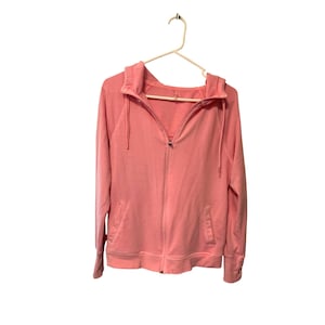 May include: A light pink zip-up hoodie with a drawstring hood and two front pockets. The hoodie is hanging on a white hanger against a white background. The sleeves have ribbed cuffs.