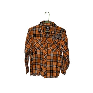 Op de afbeelding: Oranje en zwart geruit button-down shirt met lange mouwen. De shirt heeft twee borstzakken en een kleine patch op de linkerbovenborst. De shirt hangt aan een groene hanger.
