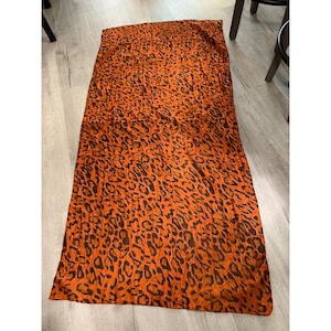 May include: An orange scarf with a brown leopard print pattern. The scarf is rectangular and appears to be made of a lightweight fabric. The leopard print is evenly distributed across the scarf.
