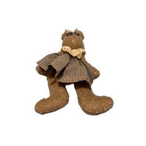 Könnte beinhalten: Ein brauner Teddybär, der ein kariertes Kleid mit einer Spitzenborte und einer Schleife auf dem Kopf trägt.