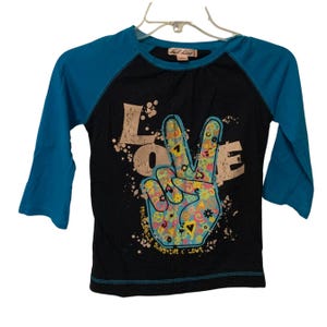 Puede incluir: Camiseta negra de estilo raglán con mangas turquesas. La parte delantera presenta un colorido signo de la paz y la palabra "LOVE" en una fuente desgastada. El texto adicional incluye "PEACEABLE SUNSHINE & LOVE".