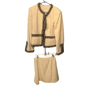 May include: Cream-colored tweed skirt suit with black trim. The jacket has two front pockets and a black trim around the collar, front, and cuffs. The skirt is a matching cream tweed. The suit is hanging on a hanger.