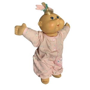May include: A Cabbage Patch Kid doll wearing a pink and white patterned sleeper with a heart-shaped patch. The doll has blonde hair and blue eyes.