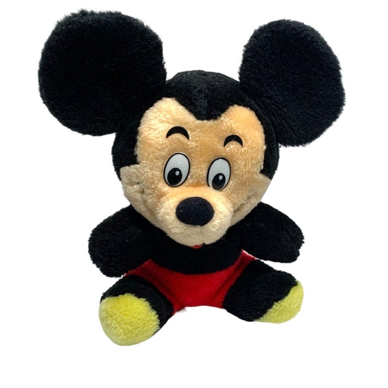 Może przedstawiać: Pluszowa zabawka Mickey Mouse siedząca z założonymi nogami. Ma czarne uszy, czarny nos i ż&oacute;łte buty. Jego twarz jest brązowa i nosi czerwone spodenki.