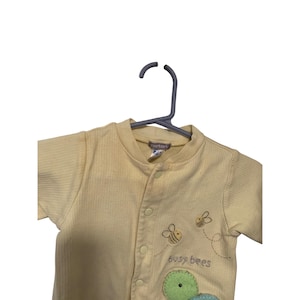 Pu&ograve; includere: Un body per neonati giallo chiaro con una trama a coste, con ricami di api e la scritta "busy bees". Ha un colletto rotondo e chiusure a scatto sul davanti. L'etichetta del marchio Carter's &egrave; visibile.