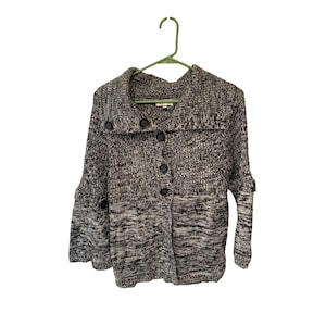 Può includere: Un cardigan con bottoni in maglia marrone e grigio screziato. Il maglione ha un ampio colletto, maniche lunghe e grandi bottoni scuri. È appeso a una gruccia verde su sfondo bianco.