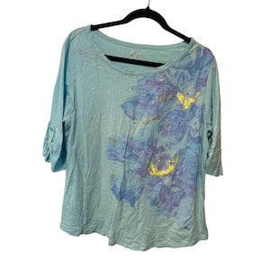 Puede incluir: Camiseta azul claro de manga tres cuartos con diseño floral. La camiseta tiene cuello redondo y mangas enrolladas. El diseño floral es en tonos azules, morados y amarillos.