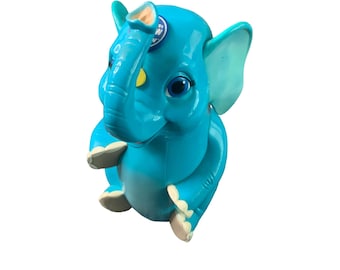 Può includere: Un elefante giocattolo di plastica blu con un adesivo giallo e bianco sulla fronte. L'elefante ha una lunga proboscide e grandi orecchie.