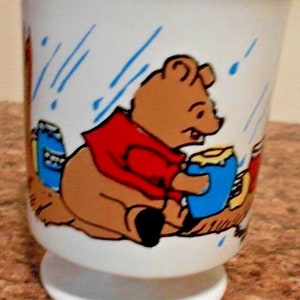 May include: White ceramic mug featuring a Winnie the Pooh illustration. Pooh Bear is wearing a red shirt and holding a blue jar of honey. The mug has a blue and white rain design.