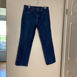 May include: A pair of blue denim jeans hanging on a hanger against a white wall.
