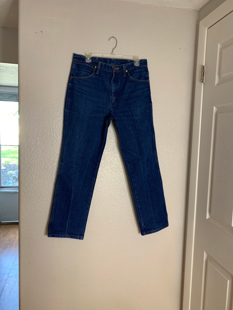 May include: A pair of blue denim jeans hanging on a hanger against a white wall.
