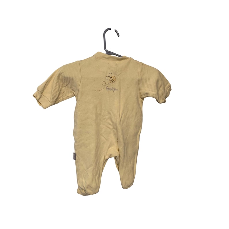 Pu&ograve; includere: Un pigiama per neonati giallo chiaro con maniche lunghe e piedini. Il retro del pigiama presenta un'ape ricamata e la parola "busy". Il pigiama &egrave; appeso a un appendiabiti nero.