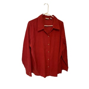 Puede incluir: Una camisa de manga larga con botones en un tono rojo intenso. La camisa presenta un cuello y está colgada de una percha blanca. La tela tiene una textura sutil y los botones son de un tono complementario.