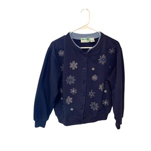 May include: Navy blue button-up sweatshirt with snowflake embroidery. The sweatshirt has a light blue collar and features silver and blue snowflake designs. The buttons are navy blue. The sweatshirt is hanging on a white hanger.