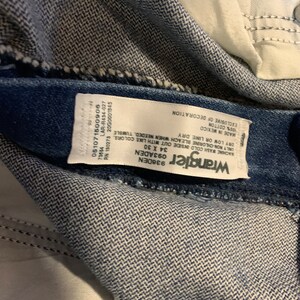 May include: A white Wrangler brand tag sewn into a pair of blue denim jeans. The tag includes care instructions, size, and material information. The text on the tag reads: "Wrangler 936DEN 093ADEN 34 X 30 051071500906 73654 RN 130273 2000001845 LAR-R434-027 WACHINE WASH COLD INSIDE OUT WITH LIKE COLORS ONLY NON-CHLORINE BLEACH WHEN NEEDED, TUMBLE DRY LOW OR LINE DRY MADE IN MEXICO 100% COTTON EXCLUSIVE OF DECORATION"