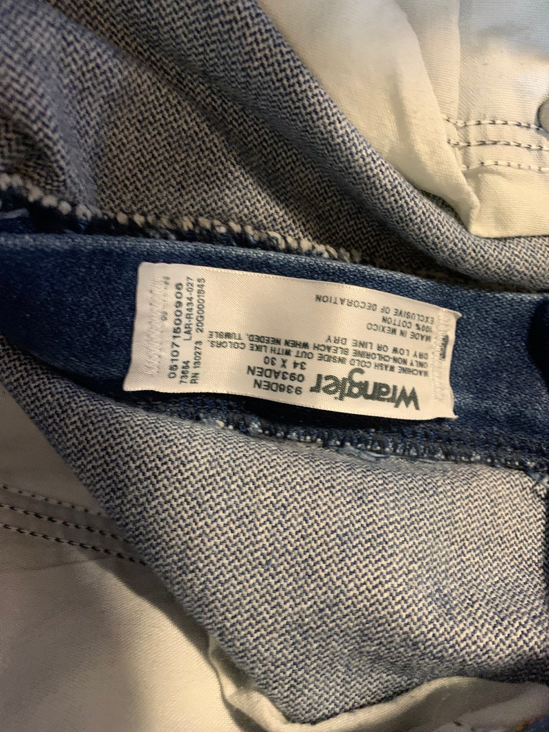 May include: A white Wrangler brand tag sewn into a pair of blue denim jeans. The tag includes care instructions, size, and material information. The text on the tag reads: "Wrangler 936DEN 093ADEN 34 X 30 051071500906 73654 RN 130273 2000001845 LAR-R434-027 WACHINE WASH COLD INSIDE OUT WITH LIKE COLORS ONLY NON-CHLORINE BLEACH WHEN NEEDED, TUMBLE DRY LOW OR LINE DRY MADE IN MEXICO 100% COTTON EXCLUSIVE OF DECORATION"