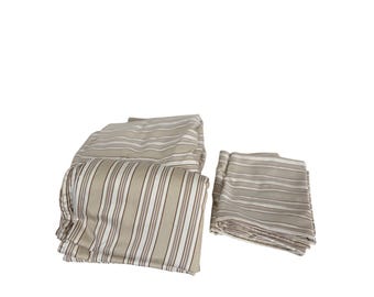 Può includere: Un set di biancheria da letto piegata caratterizzato da un motivo a righe verticali nei toni del beige, bianco e marrone scuro. Il tessuto appare liscio ed è disposto in due pile distinte, che mostrano il design a righe uniforme.