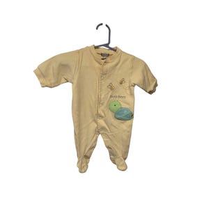 Pu&ograve; includere: Un pigiama per neonati giallo chiaro con maniche lunghe e piedini attaccati, appeso a una gruccia nera. Il pigiama ha un'abbottonatura frontale e presenta un disegno ricamato di una tartaruga verde e le parole "busy bees".